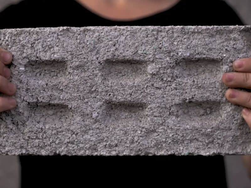 A man holding up a plastic-based brick with two hands. The brick has six indentations in its face and is light gray with a granular texture. The man holding the brick is wearing a black T-shirt.