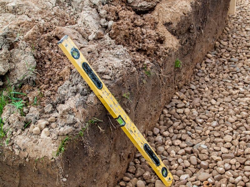 A trench filled with large stones to serve as a foundation for an earthbag house. There is a yellow spirit level leaning on the side of the trench.