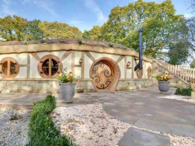 A half-timbered hobbit house made from earthbags. It is buff-colored and has light brown timbers. The door and windows are round and the door has ornate patterns on it. The area outside is paved with stone slabs and there are plant pots either side of the entrance.