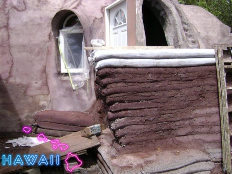 A photograph of an earthbag house under construction. there is a wall in the foreground that has the layers of earthbag still clearly visible and the facade behind is covered with plaster and has white doors and windows fitted. In the bottom left is a stylized map of Hawaii with the outline of the islands in pink and the name "Hawaii" in blue lettering.