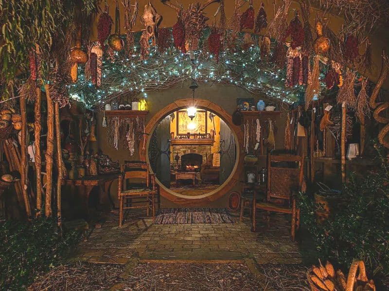 A beautiful hobbit house with a circular front door, beyond which can be seen the fireplace and mantelpiece. There is amazing attention to detail in recreating the hobbit house, with tools, utensils and decorations handing from the walls. There are two seats in the porch, which is paved with stone and has a mat at the doorway.