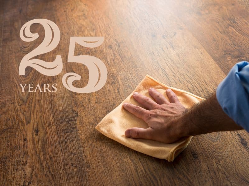 A photograph of a man's hand wiping a shiny brown engineered hardwood floor with a beige-colored cloth. The floor has a beautiful grain and the top left corner of the image has "25 years" written in it in an ornate beige font.