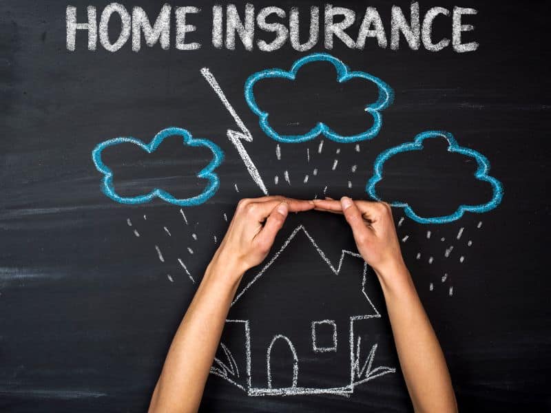 A blackboard and chalk drawing of a house in a storm with rain falling from clouds and lightning. The words "home insurance" are written along the top of the blackboard and a person has their hands held up in front of the drawing so they appear to be protecting the house from the weather.