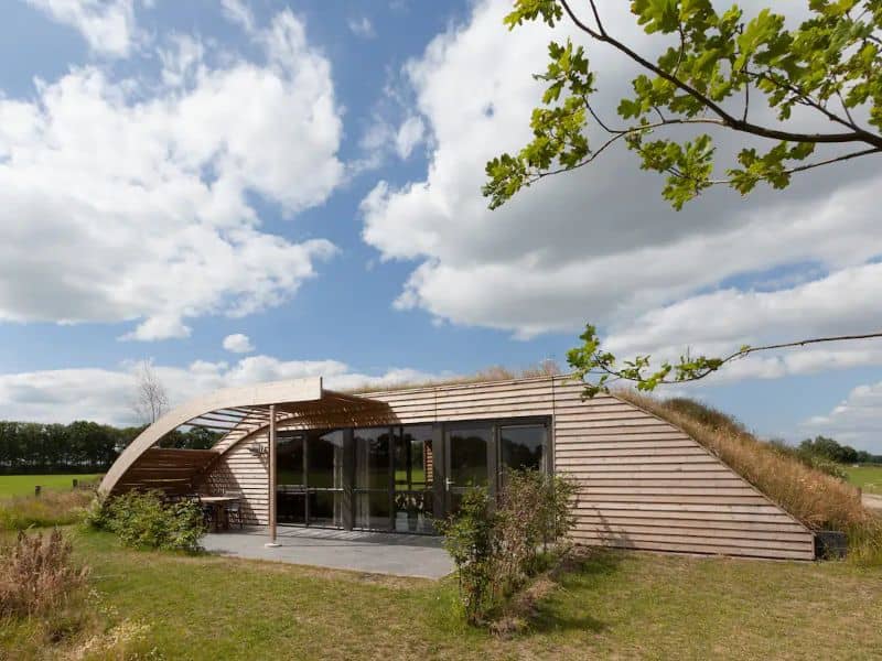 An earthbag house with a domed roof that has grass growing on top of it. The front wall is clad with wooden planks and has large, full-length windows and doors. There is a curved pergola attached which provides cover to the paved patio area out front. Beyond the plot are fields and trees.