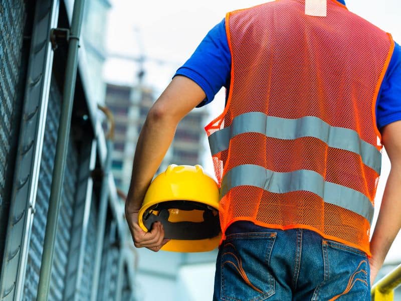 A construction worker walking away from the camera holding a yellow hard hat on his hip. He is wearing blue jeans and an orange high visibility tabard. On the left of the photo is a metal fence and an out of focus high-rise building with a crane on top is in the background.