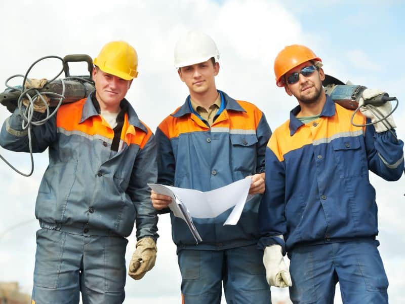 Three construction workers in overalls and hardhats. Two are holding power tools and one has a set of plans.