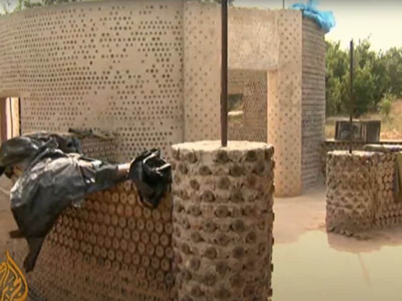 A screenshot from an 
Al Jazeera English video on YouTube. The video covers the initiative in Nigeria to use plastic waste for construction of houses. The screenshot shows a plastic bottle house made using water bottles filled with compacted dirt and bound with mud and string. The walls are pleasantly curved and the main house is round. The ends of the water bottles can clearly be seen in the walls. There is protective plastic sheeting covering the tops of the walls as the house is still under construction.