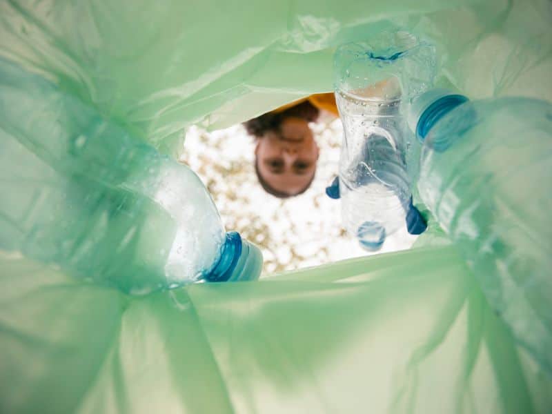 The collection of plastic water bottles for recycling. The photograph is taken from inside the collection bag, which is translucent and green. The plastic bottles in the bag are clear with a slight blue tinge. The person holding the bag is seen through the neck of the bag as the camera is located in the bottom of the bag looking up.