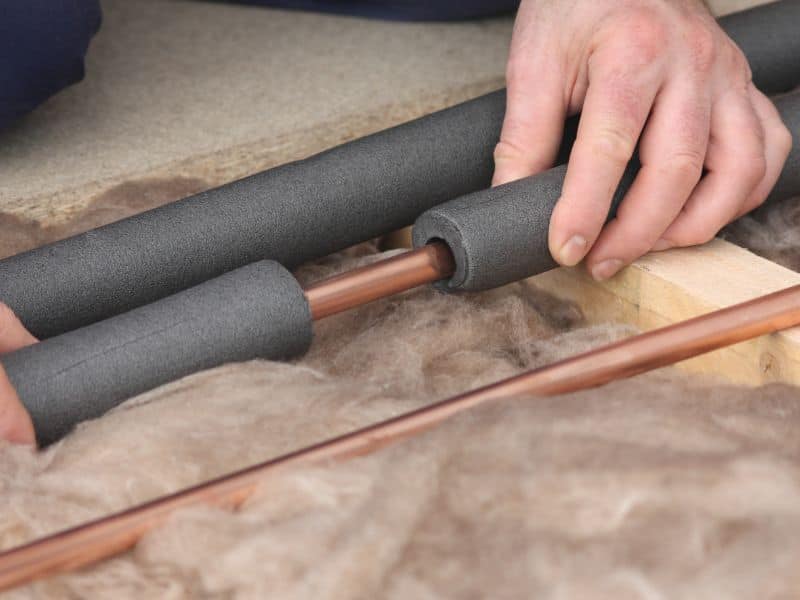 A plumber placing recycled plastic foam insulation around a copper pipe in a floor void over the top of rockwool.