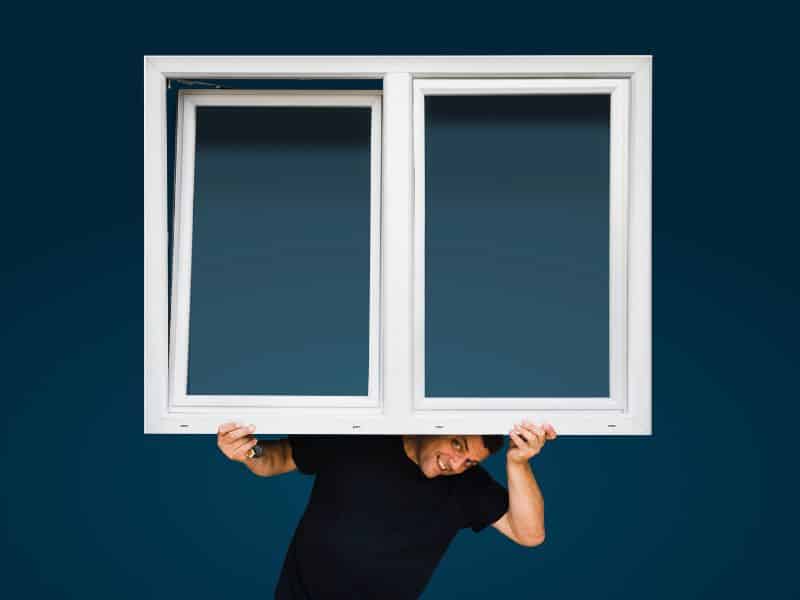 A man carrying a white plastic window frame in front of a blue background. There are two panes, the left one is ajar. The man is wearing a blue T-shirt and is peering out from underneath the frame with a grin on his face.