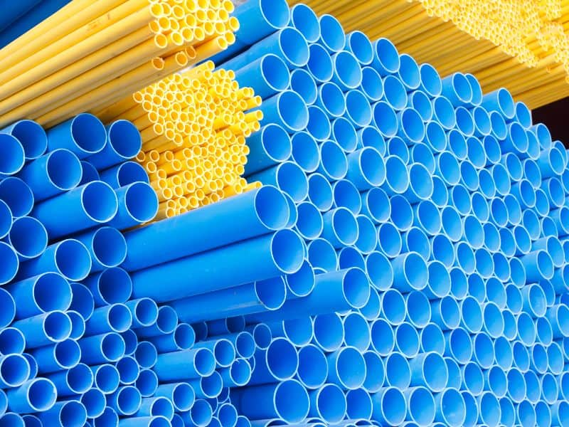 A large stack of Polyvinyl Chloride (PVC) pipes made from recycled plastic. There are large and medium diameter blue pipes and smaller bore yellow pipes. The different colored pipes are grouped together.