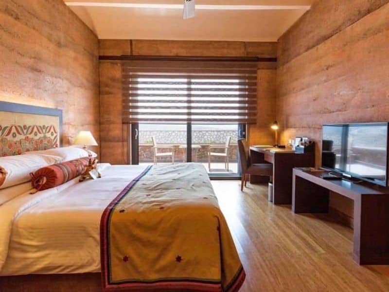 A bedroom with rammed earth walls and a polished wooden floor. There is a large double bed in the room, a desk and a dressing table with chairs. The end wall has large glass doors leading onto a balcony.