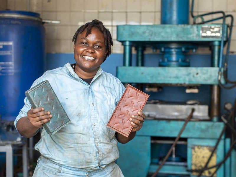 A photograph of Nzambi Matee, founder of startup company Gjenge Makers holding two of the recycled plastic bricks invented and manufactured by her company. She is wearing light-blue work clothes and there is a hydraulic press in the background that is used to make the bricks.