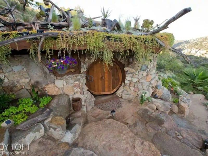 An earthbag hobbit house built into the hillside. The walls are clad with stone and there is natural stone paving outside. The wooden door is natural wood and circular in shape. There are tree branches arranged on top of the house.