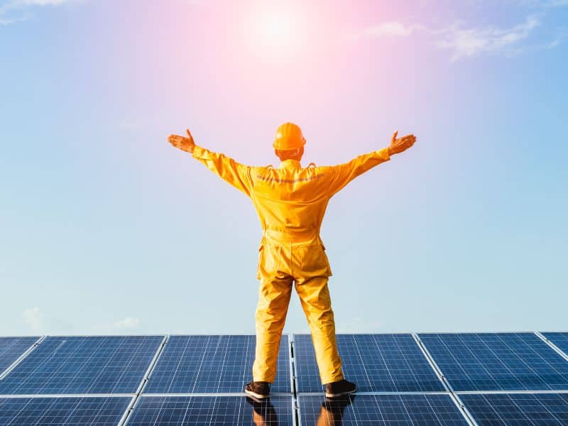 A photograph of a man dressed in yellow overalls and a yellow hardhat with his back to the camera and facing the sun with arms outstretched. He is standing on an array of solar panels.