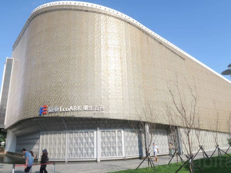 The EcoArk in Taipei. It is made from recycled plastic bottles, has nine stories and a sweeping curved facade. It is a light beige color and has a logo saying "EcoARK" on the front. The ground floor has diamond-shaped corrugations along it and there is a paved area in front with people walking by.