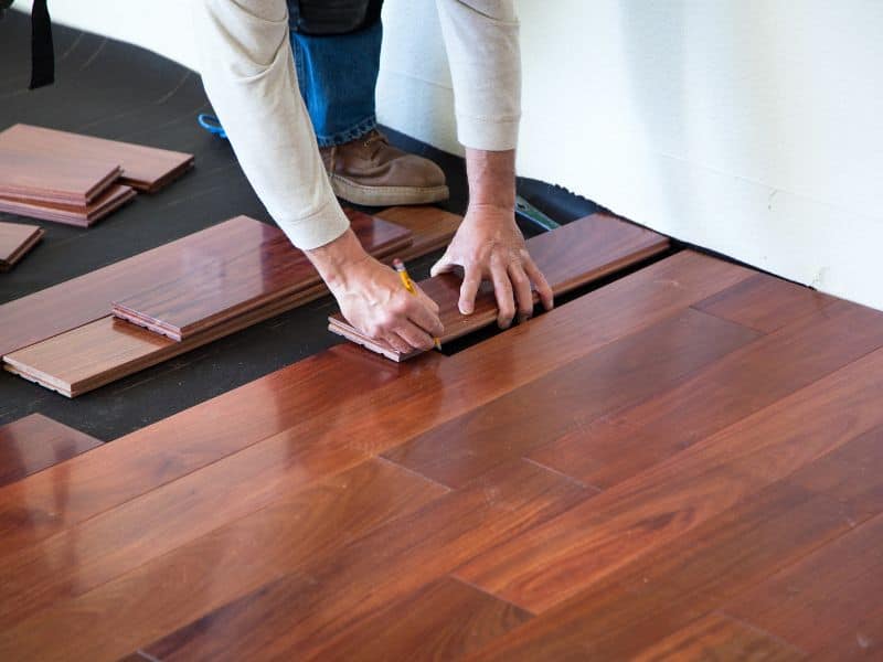 A hardwood floor being laid by a worker who is marking a piece of wood before cutting it to size. The wood is a rich dark brown color and has a high shine finish. The black underlay for the wooden floor is visible on the unfinished section of floor.