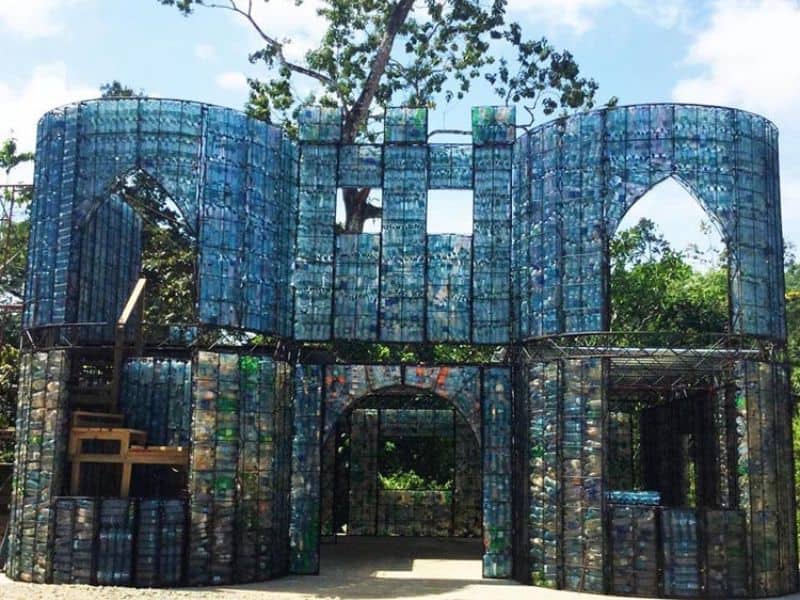 A photograph of a two-story building under construction. The walls are made from metal cages filled with waste plastic bottles. The holes for the windows are small squares, round arches and gothic arches, giving it a somewhat eastern appearance.