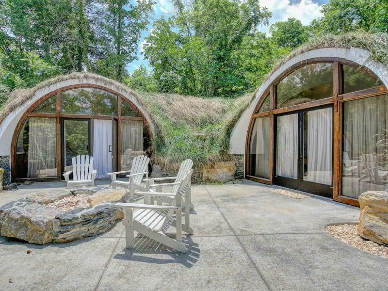 An earthbag house comprising two domes with their gable ends containing floor-to-ceiling glass windows and doors with drapes across them for privacy. The roofs of the domes have grass growing on them to resemble small hills. There is a courtyard paved with large stone slabs and wooden deck chairs.