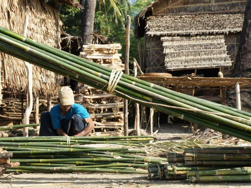 A worker crouching down next to bundles of bamboo, tying them together using natural fibers. The bamboo culms are green and the lashings he is using are light yellow. There are bamboo buildings in the background.