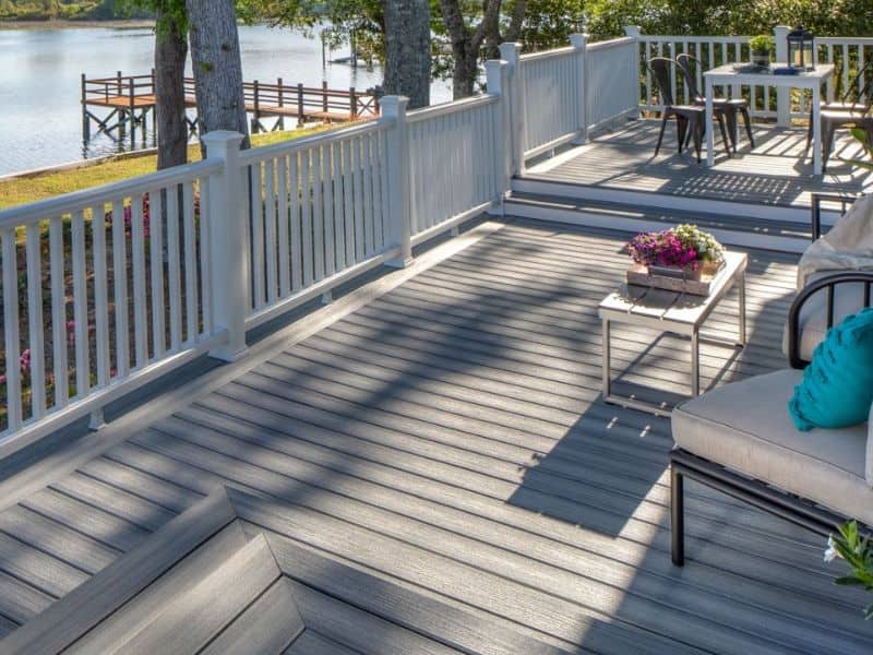 Trex Enhance decking in a beautiful natural wood-like finish. There is a white railing around the edge and patio furniture with black metal frames and cream-colored cushions. There is a lake with a jetty and trees in the background.