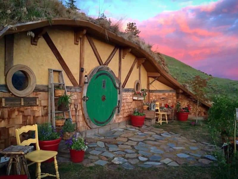 An earthbag house built into the hillside. It has a round green door in the front and timbers in the wall to support the roof overhang. There is crazy paving out front and a couple of yellow wooden chairs.