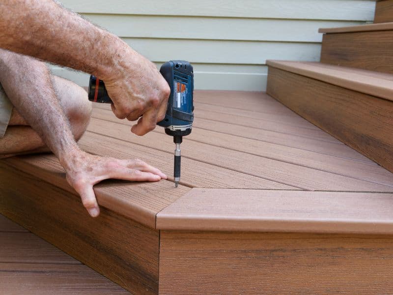 A composite deck being installed. The image shows a worker using a drill to drive screws into the first step leading up a series of steps. The composite wood is a rich brown color and looks similar to natural wood.