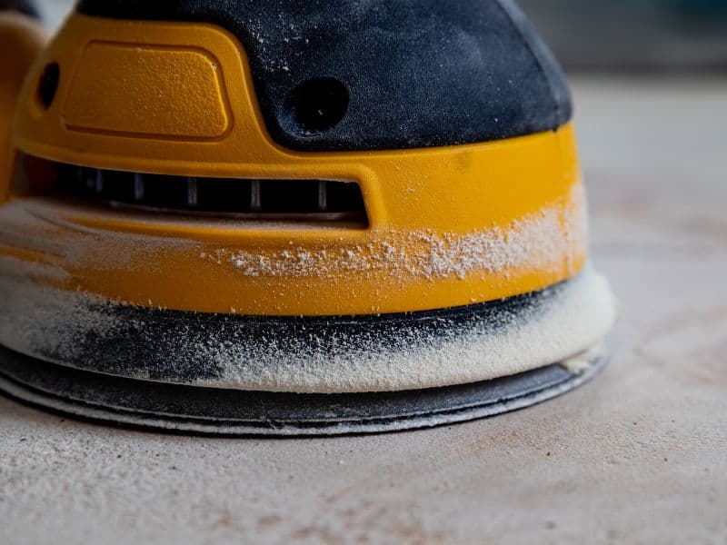 A piece of composite decking undergoing final sanding to give it a good finish. The sander is electrically-powered and yellow and black in color.