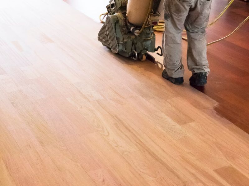 A worker sands a hardwood floor with a push-along sander with a large bag to collect the sawdust. The surface layer of the wood and polish has been removed from half of the floor, exposing the natural, clean wood below.