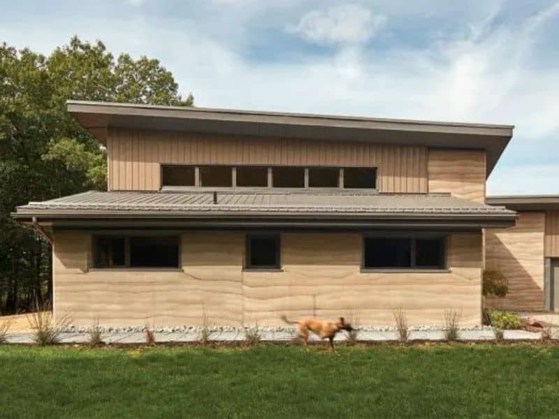 A modern, stylish, light brown rammed earth home with small windows and a metal roof. There are two dogs running across the lawn in front of the building.