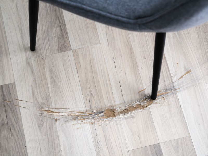 A light brown engineered wood floor has been damaged by a sharp metal chair leg being repeatedly dragged across it.