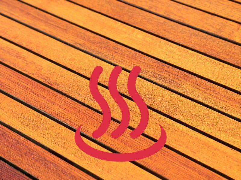 A closeup photograph of a decking area in mid-brown composite wood-effect decking boards. The boards run diagonally across the image from top left to bottom right. There is a "hot" icon overlain which comprises red lines with a single curve in a smile shape with three vertical wavy red lines above that resemble steam rising.