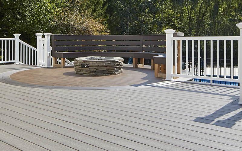 A photograph of TimberTech decking in direct sunlight. There is a stone fireplace structure with seating around it and white railings. The decking boards are light gray, which helps reflect the sun's heat.