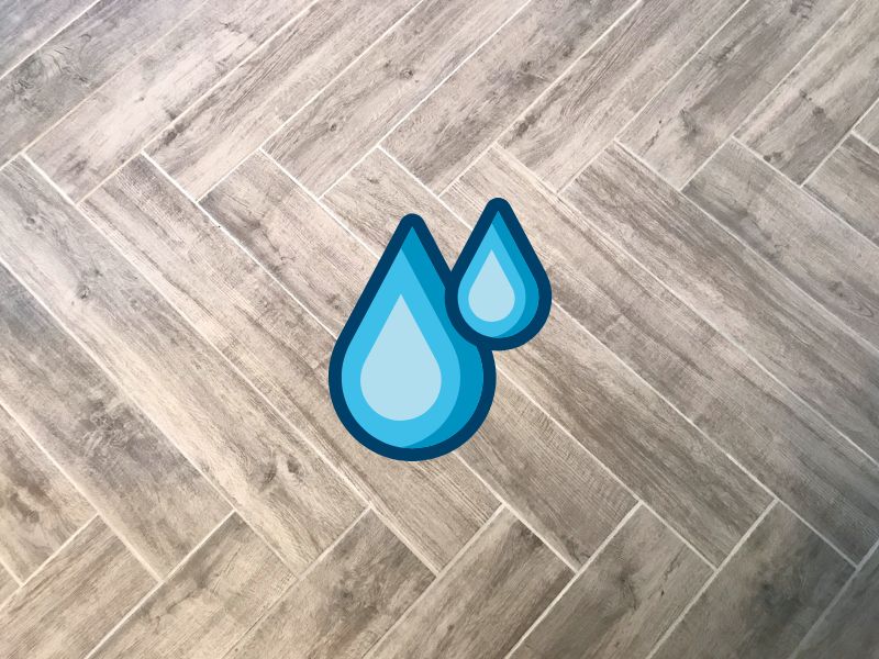 A light brown herringbone-patterned engineered hardwood floor with two cartoon water droplets overlain.