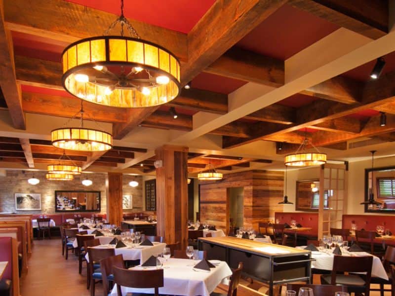 A restaurant with beautiful reclaimed wood beams. You can see the saw marks on the beams, along with grain patterns and knots. There are circular light fittings hanging from the ceiling. and the wood panels between the beams are painted red.