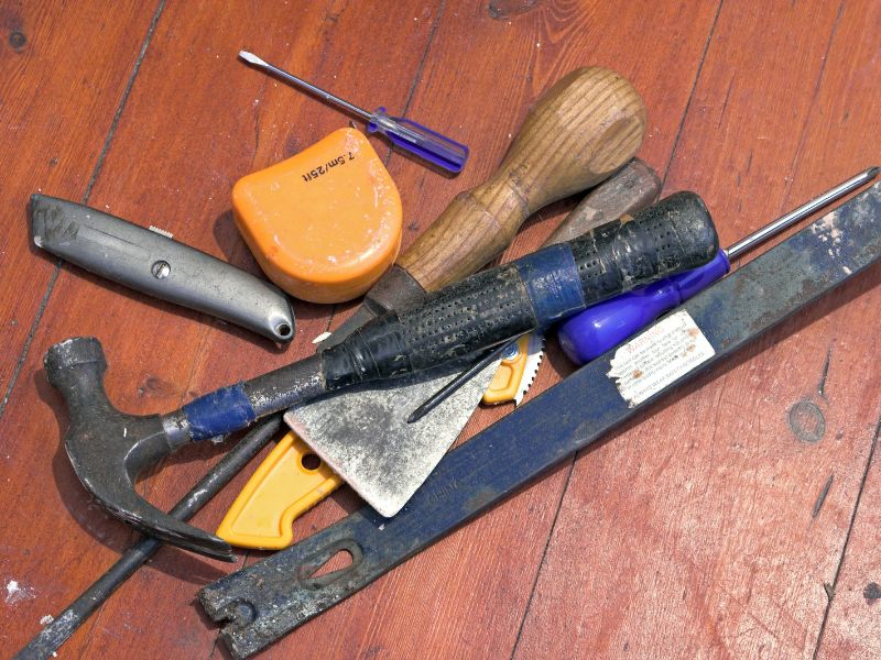 Assorted tools are laid out on a wooden floor. The tools include a measuring tape, screwdriver, hammer and utility knife.