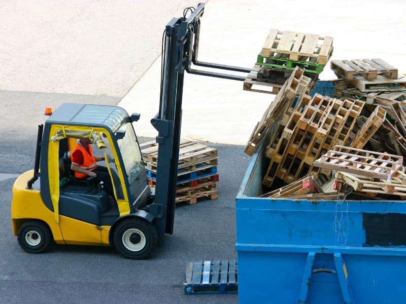 A yellow fork lift truck lifting waste wooden pallets into a blue skip.