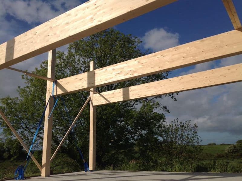 A structural frame for a large building made from long spans of glulam timber. The timbers are attached to a concrete slab and are temporarily held in position with blue straps and temporary wooden cross-pieces.