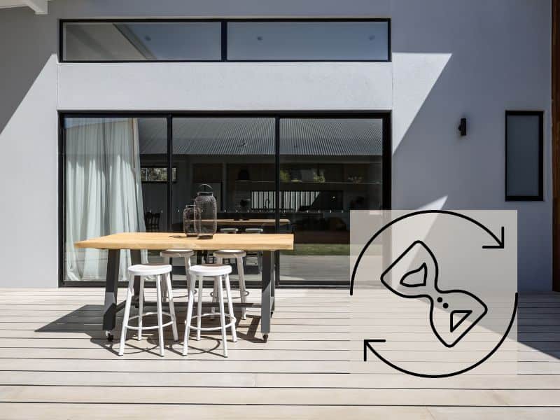 A light-colored deck outside a modern stylish house with pale gray walls and large floor-to-ceiling windows with black metal window frames and white drapes on the left side. On the deck is a table and four stools. There is bright sunshine and in the bottom right corner is a diagonally-oriented hourglass icon with circular arrows around it.