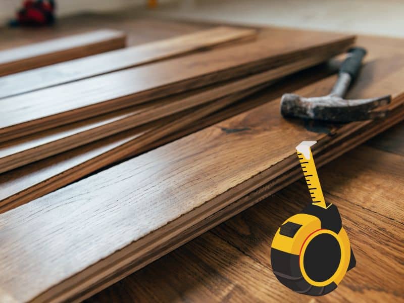 Engineered wood flooring planks stacked haphazardly over an already laid engineered wood floor. A hammer rests on top of one of the planks and a yellow-and-black cartoon tape measure is measuring the thickness of one of the planks. 