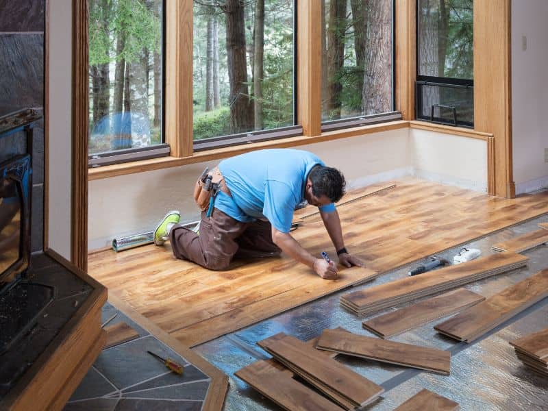 A worker installing reclaimed wood flooring in a room with a bay window. There is silver underlay visible on the floor that has yet to be covered by wood, and there are floorboards placed randomly on top of the underlay awaiting installation.