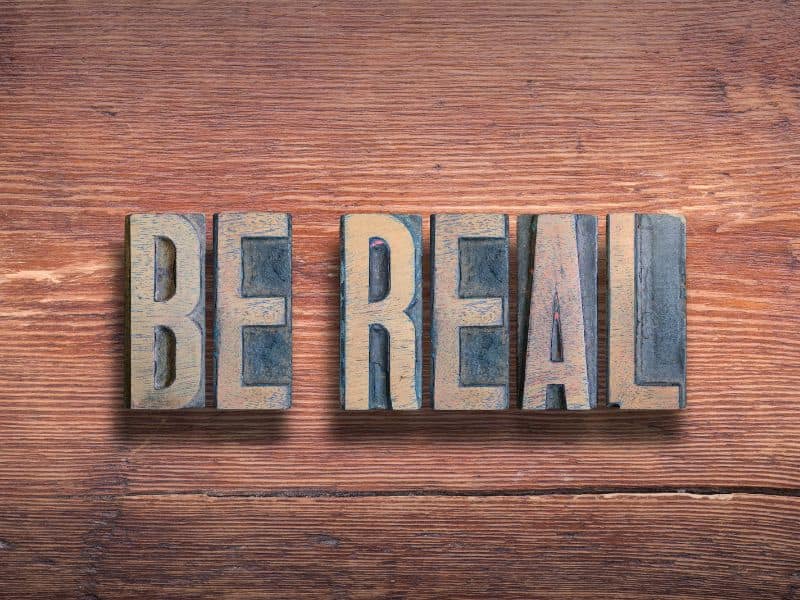 A photograph of natural dark brown wood with a visible grain and imperfections with the words "BE REAL" placed on top of it in metal blocks that look like the old "sorts" used in type-setting for printing presses.