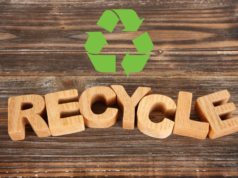 A closeup of a dark wood tabletop with wooden letters spelling the word "recycle." Above the letters is a green recycling icon with curved triangular arrows.