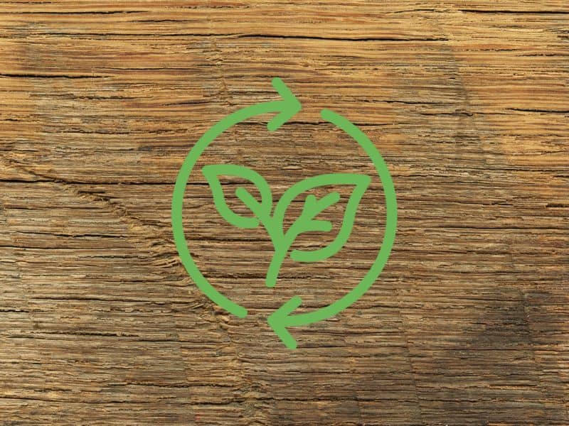 A closeup of a reclaimed wood flooring board that is quite weathered with an open, prominent grain and marks on it. Across the middle is an "eco-friendly" icon with green leaves within circular green arrows.