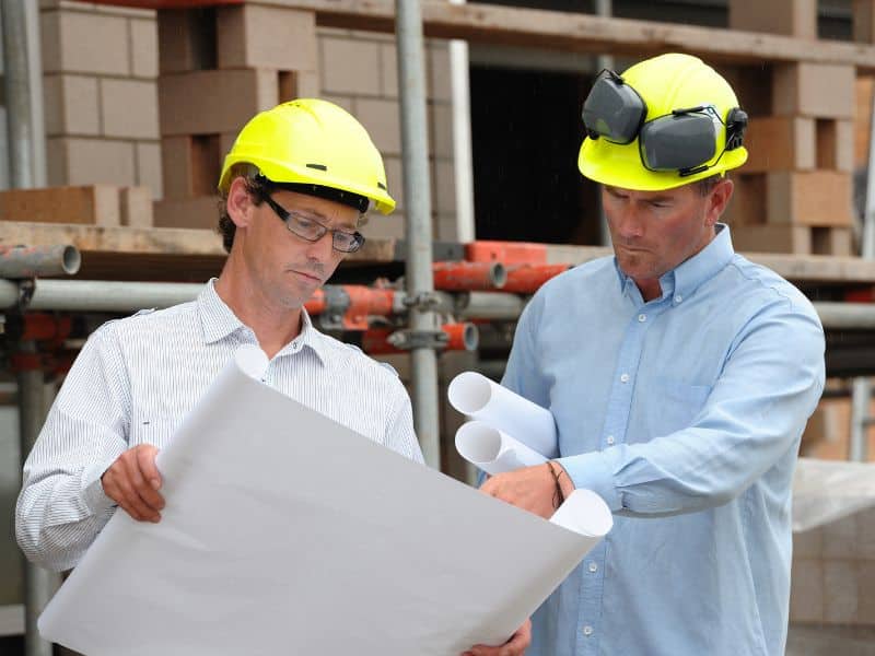 Two men in hardhats on a building site examining plans with scaffolding behind them.