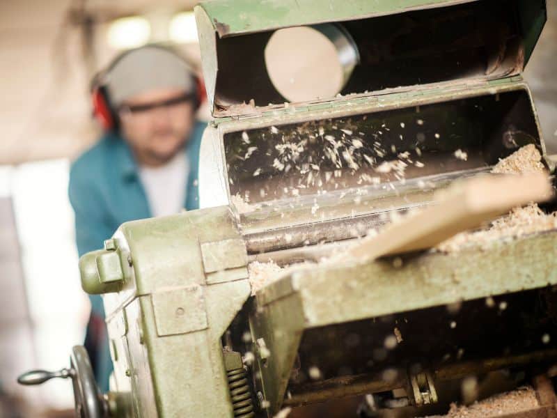 A man wearing safety glasses and ear defenders operating a wood milling machine that is removing the outer face of a wood floorboard.