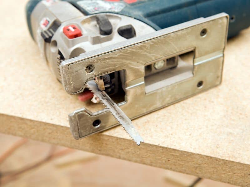 A power tool called a "jigsaw", which  is used for cutting through sheets of wood, lying on its side on top of a sheet of particleboard. The individual wood particles used in the manufacture of the particleboard are visible in the exposed cross-section along the edge of the board.