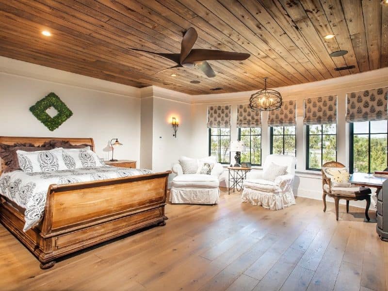 A large bedroom with a wooden ceiling and floor. The patterns in the reclaimed wood ceiling add a rustic charm to the room. Light fittings are recessed into the ceiling, and a ceiling fan hangs from the ceiling in the middle of the room.