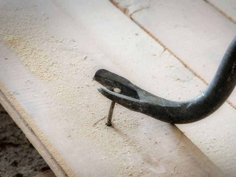 A crowbar pulling a nail out of a wood floorboard with sawdust.