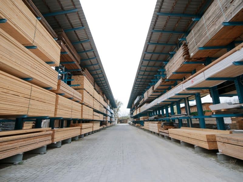 Covered shelving in a factory yard containing wood products. The photograph is taken from between two rows of shelving, looking along the aisle. The shelves are very long and are made of blue steel. The floor of the aisle is paved with Monoblock.